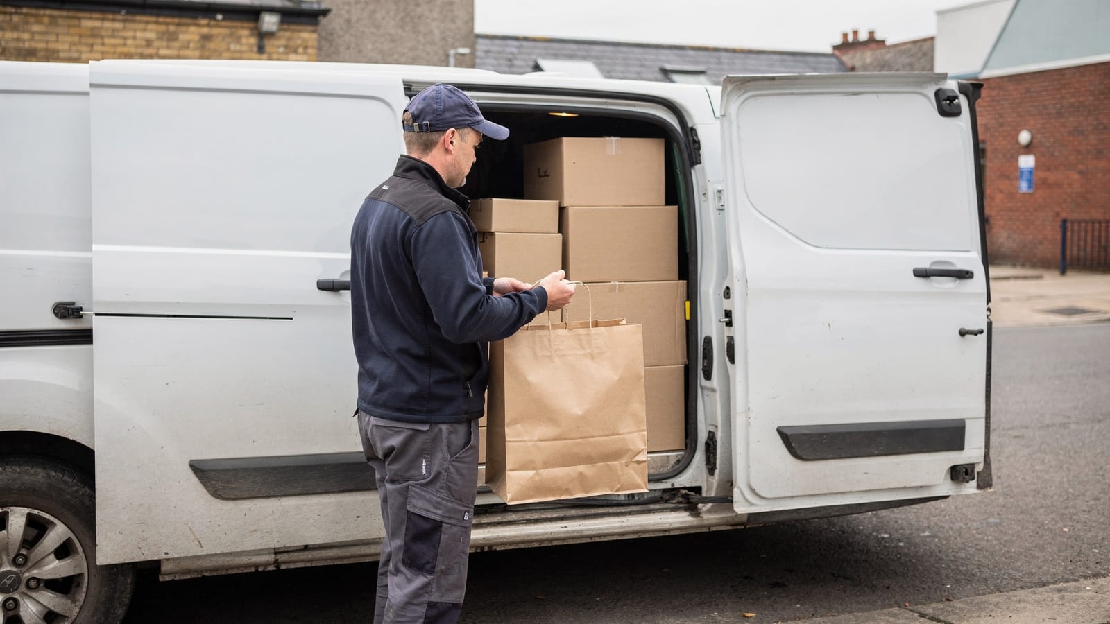A delivery driver in a cap and dark fleece holds a paper carry bag beside the open side door of a white van stacked with cardboard boxes.
