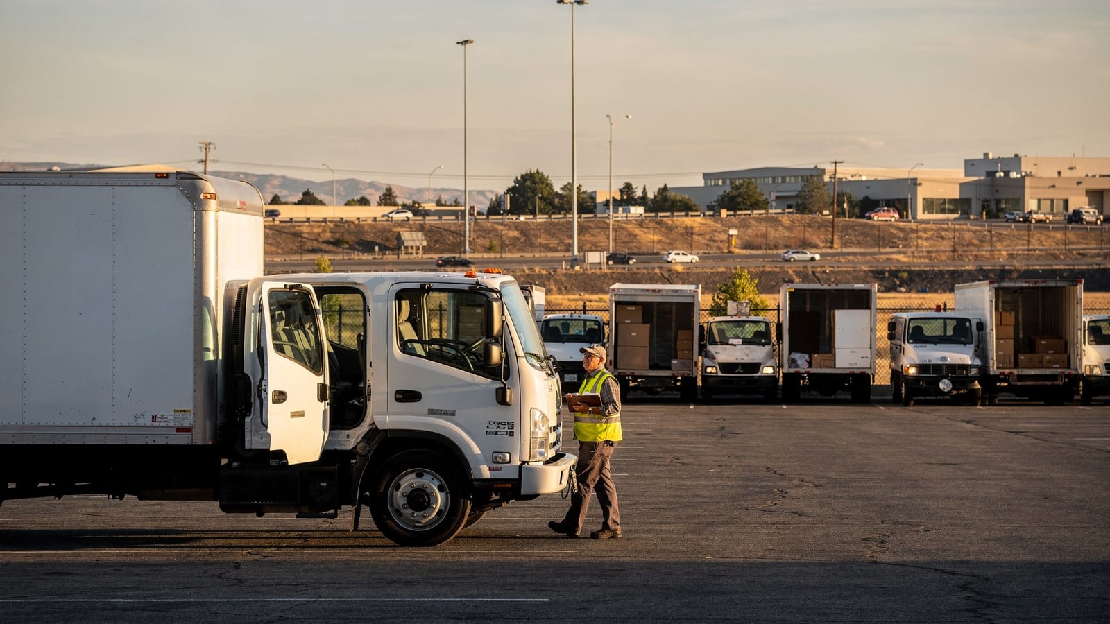 A worker in a high-visibility vest walks past a white box truck in a distribution yard at sunrise, with parked trucks and a highway beyond.